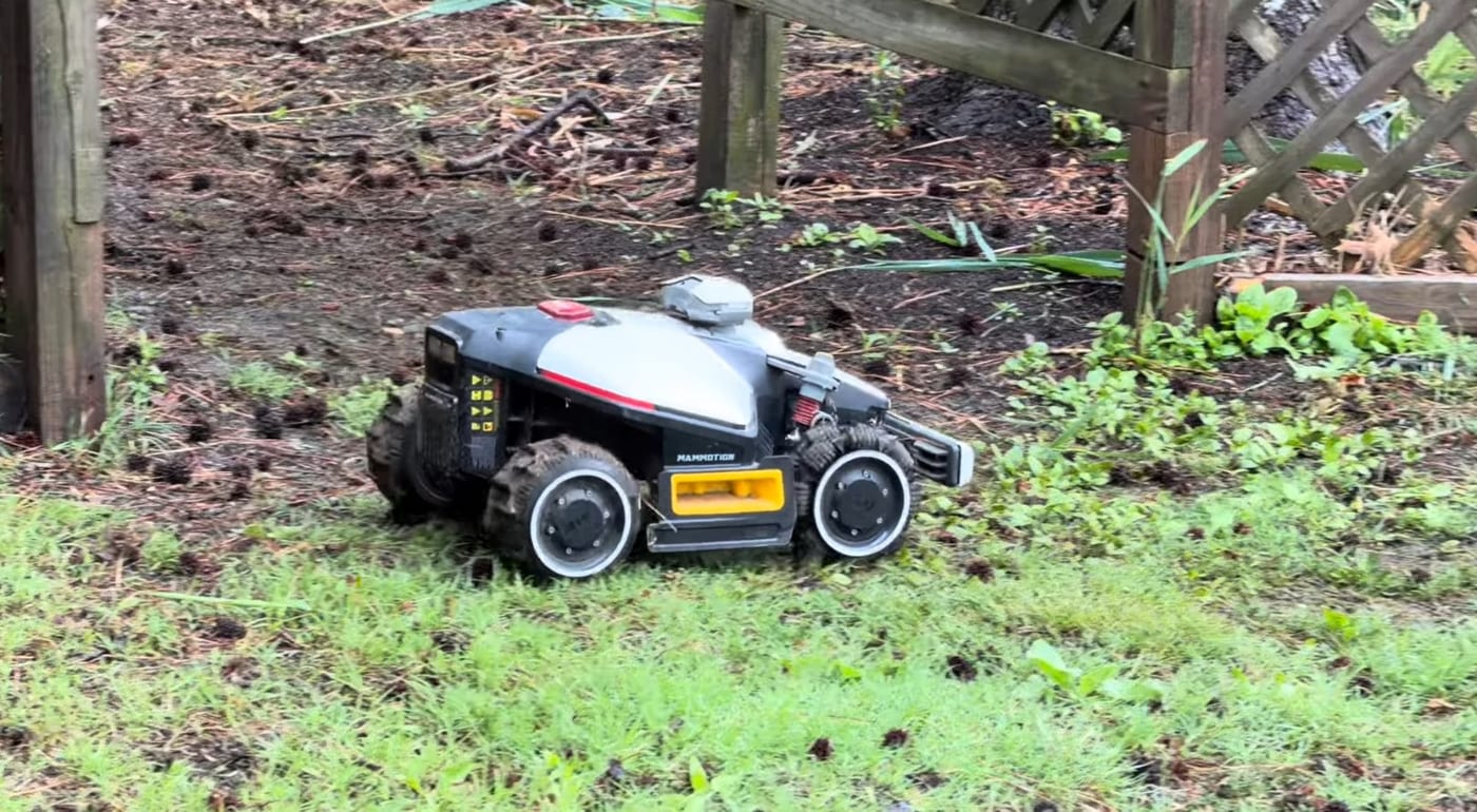 A Mammotion robot mower working in a rough, shaded patch of lawn near a fence and trellis