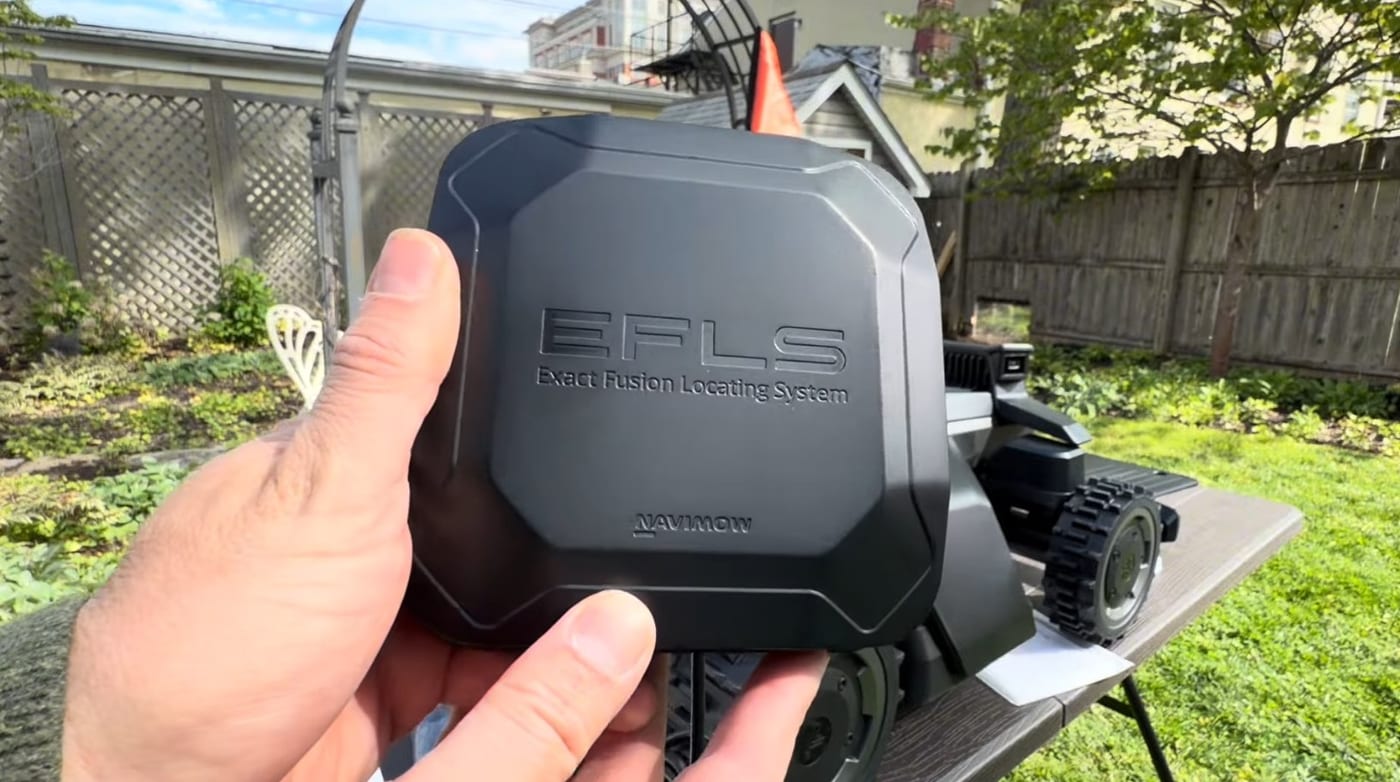 A hand holding a Segway Navimow EFLS Exact Fusion Locating System RTK receiver in front of a robot mower