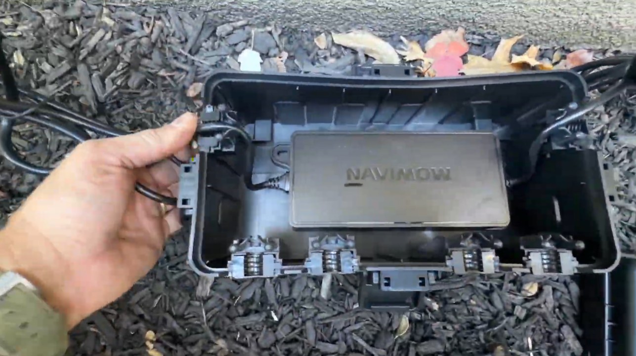 A Navimow charging station power supply inside an open outdoor enclosure box on a mulch bed, with the cables clipped through the box's grommets