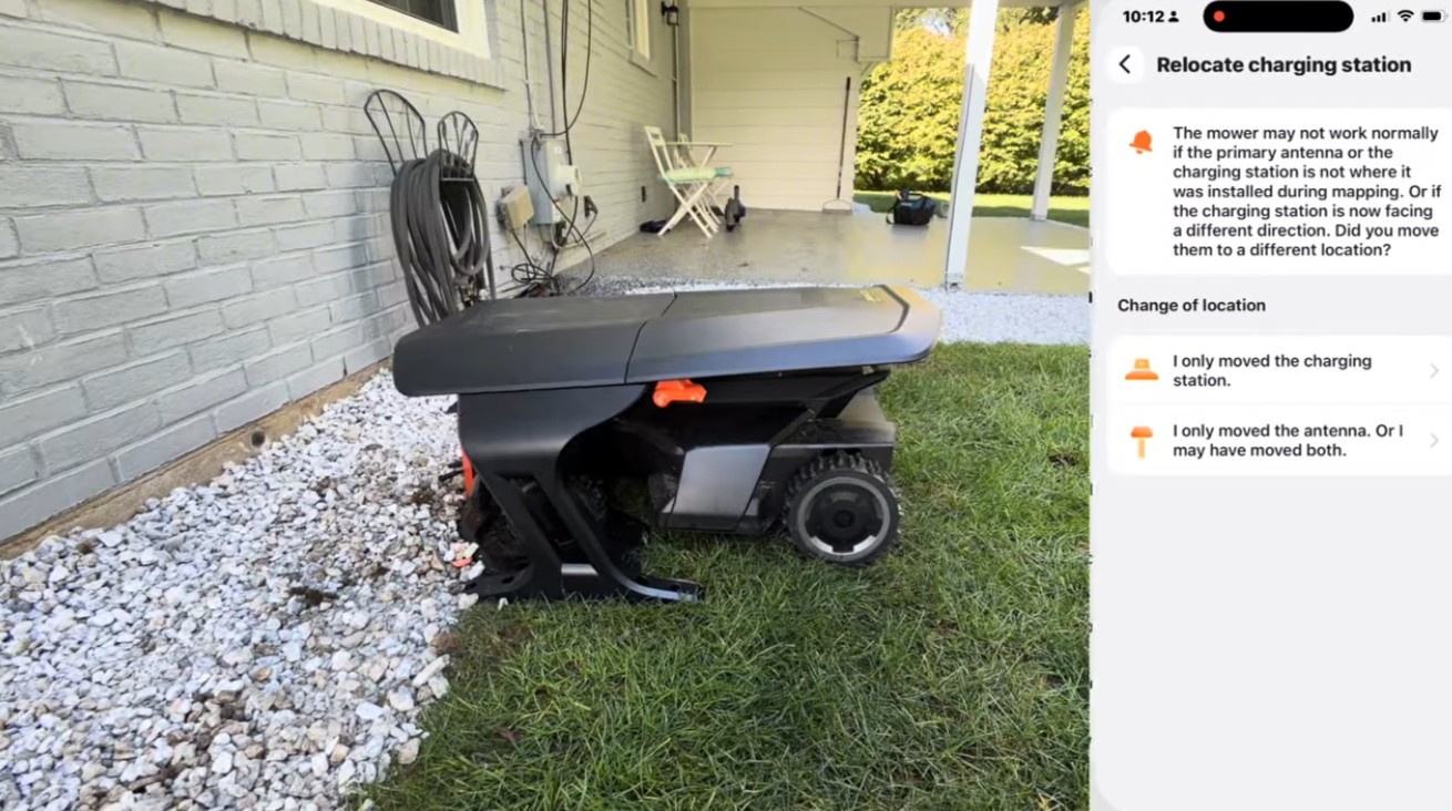 A Segway Navimow on its charging station beside a brick house wall, next to a phone showing the Navimow app's relocate charging station screen