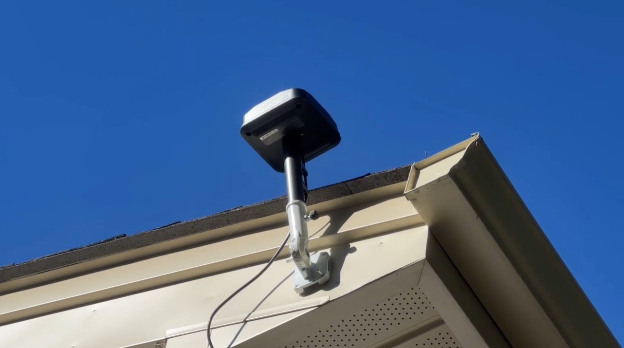 A Navimow RTK antenna on a beige wall mount at the corner of a roof eave, against a clear blue sky
