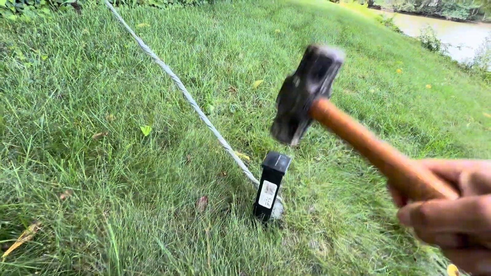 A hammer driving a stake anchor into a grass slope with tensioned steel cable running uphill