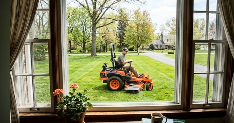 A traditional lawn service zero-turn mower cutting a front yard, seen through a living-room window