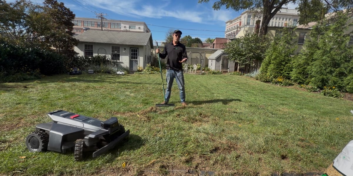 A thin backyard lawn raked out and ready for seed, a robot mower in the foreground and a bag of seed at the edge