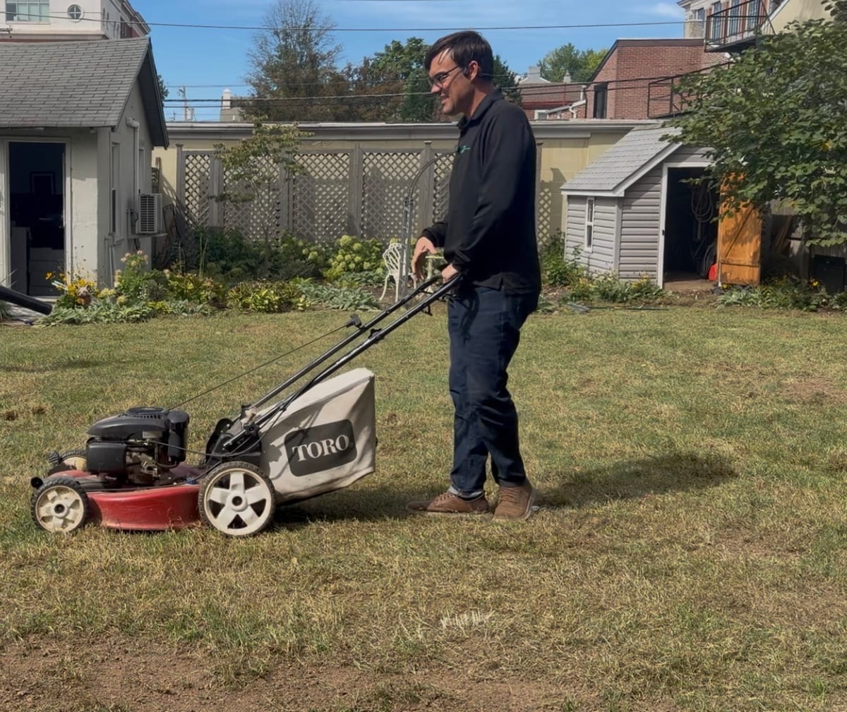 A gas push mower scalping a thin backyard lawn low before overseeding, the robot mowers parked on the edge