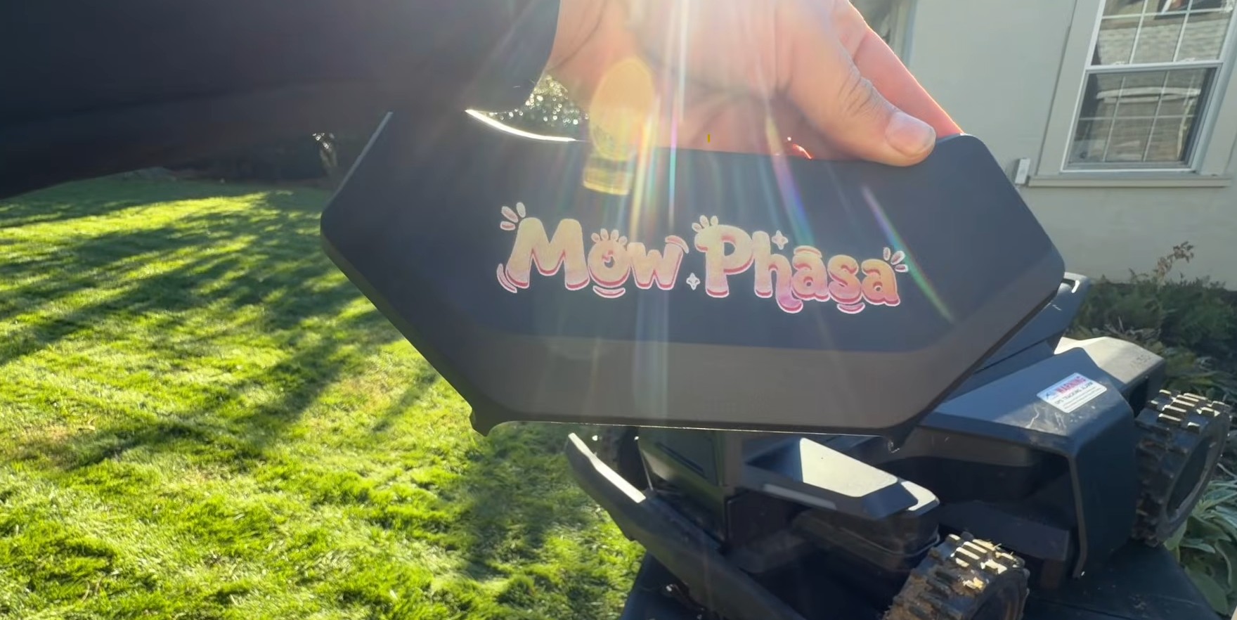 A custom Navimow nameplate reading “Mow Phasa”, UV-printed on the X4's decorative camera housing, held up in front of a freshly mowed lawn