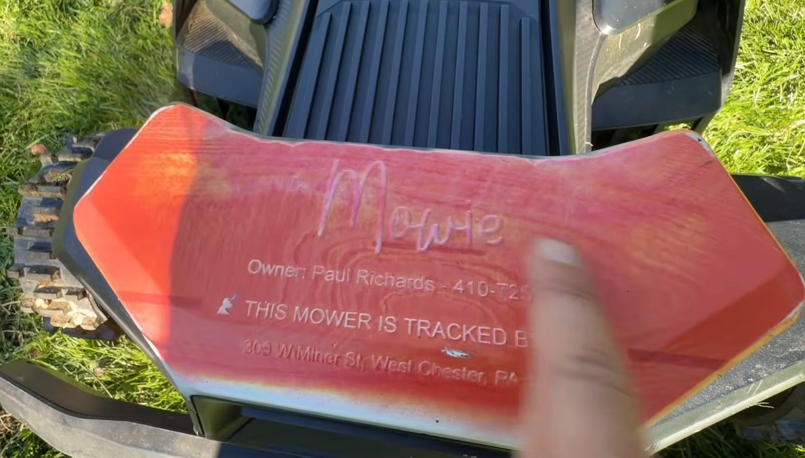 A red custom Navimow nameplate printed with the mower's name, the owner's name and phone number, and a line saying the mower is tracked